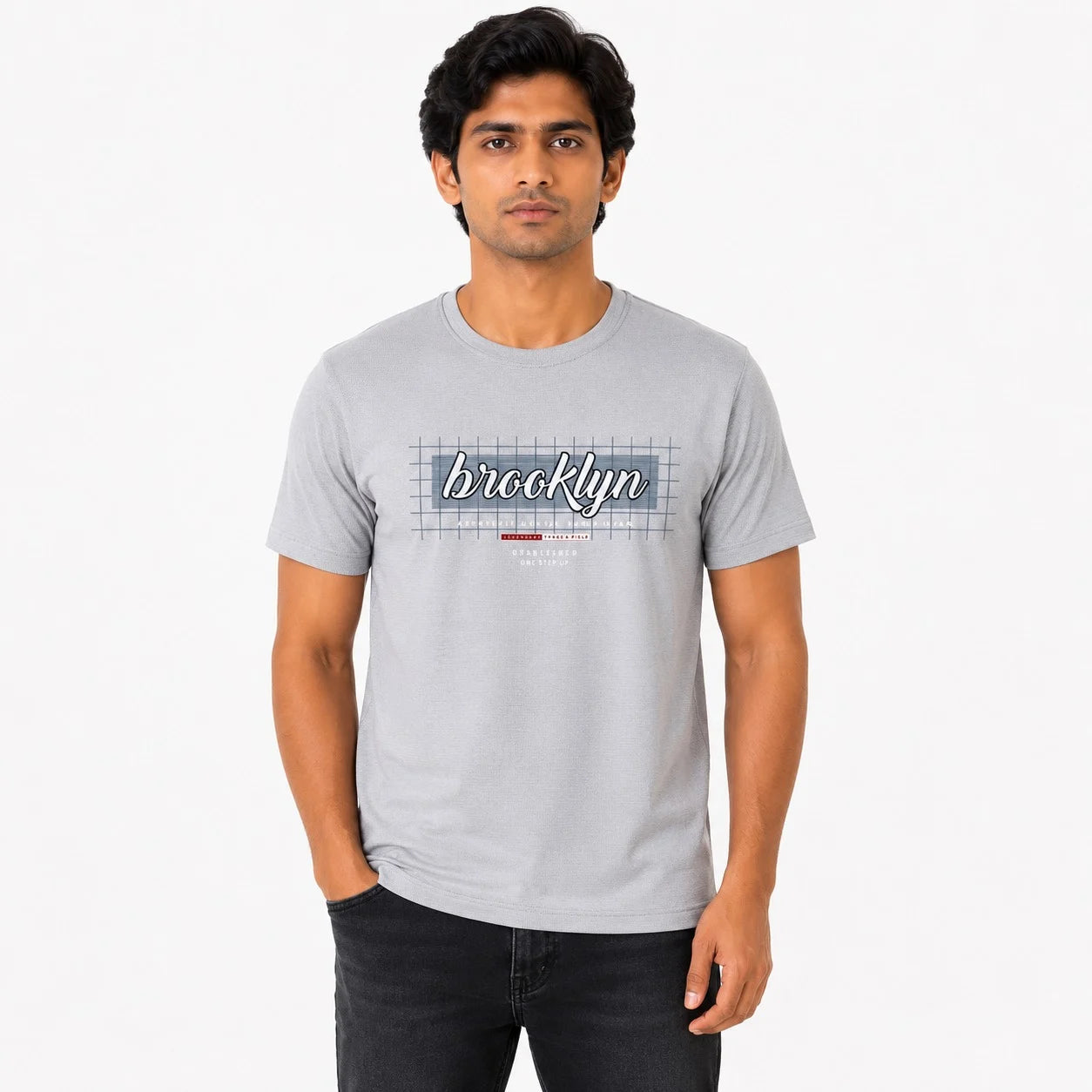 A man wearing a light gray crewneck t-shirt with a 'Brooklyn' graphic on the chest, paired with dark jeans, standing against a plain white studio background.
