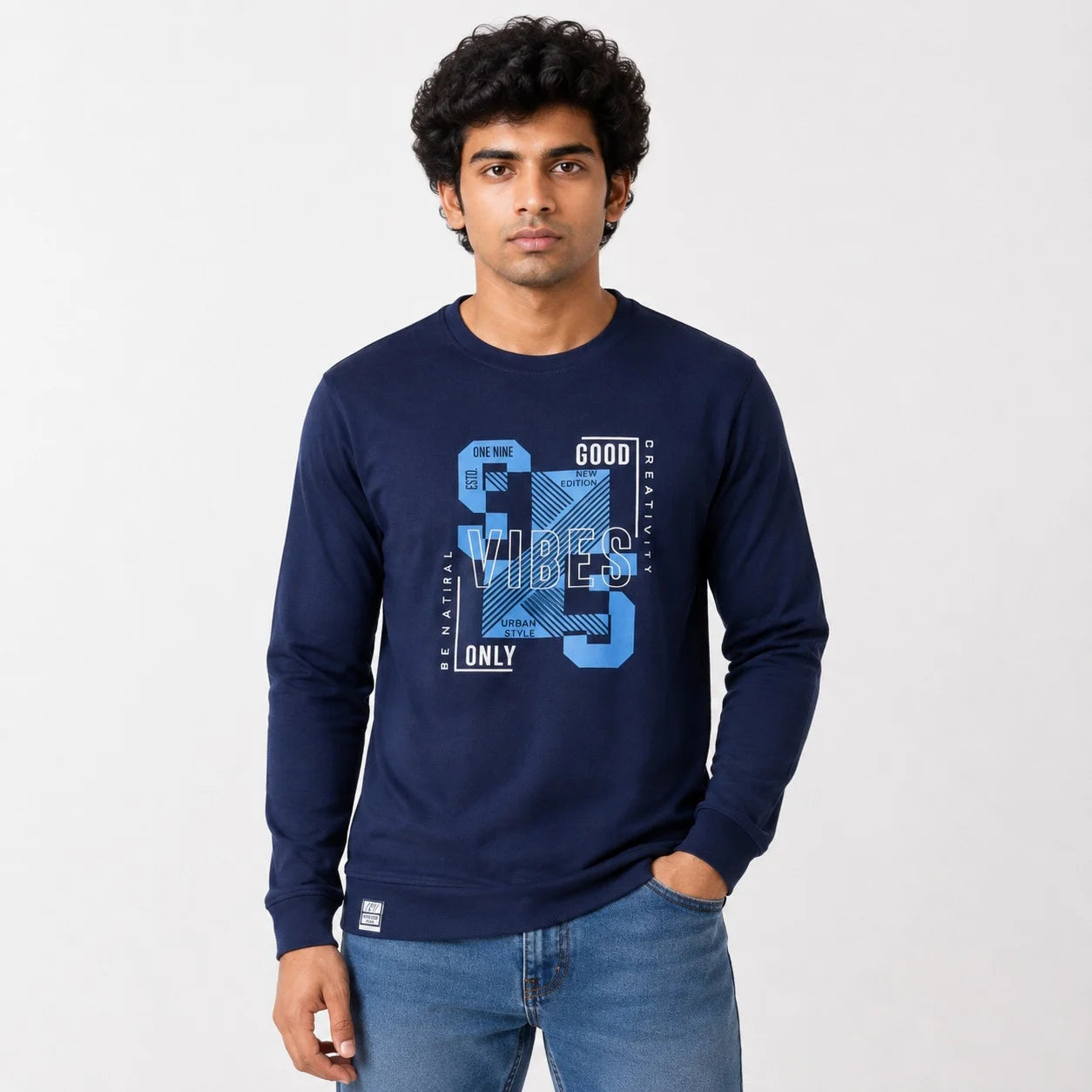A man wearing a navy blue long-sleeve t-shirt with a blue and white graphic print on the front, paired with blue jeans, standing against a light gray background.