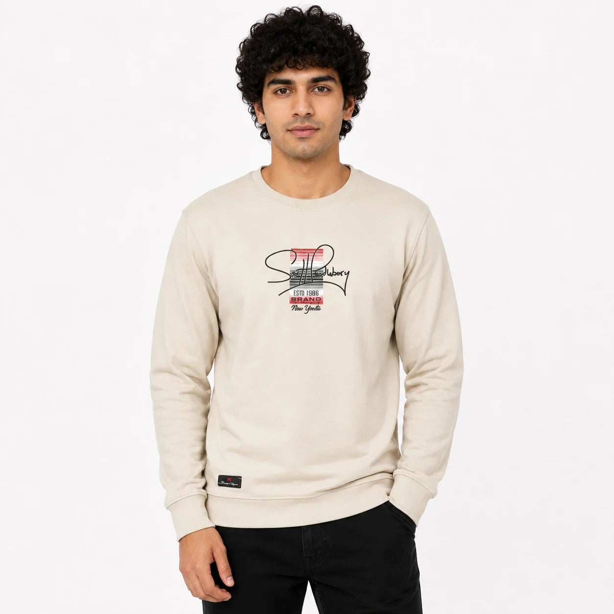 A man wearing a cream-colored crewneck sweatshirt with a graphic print on the chest, paired with black pants, against a plain white background.