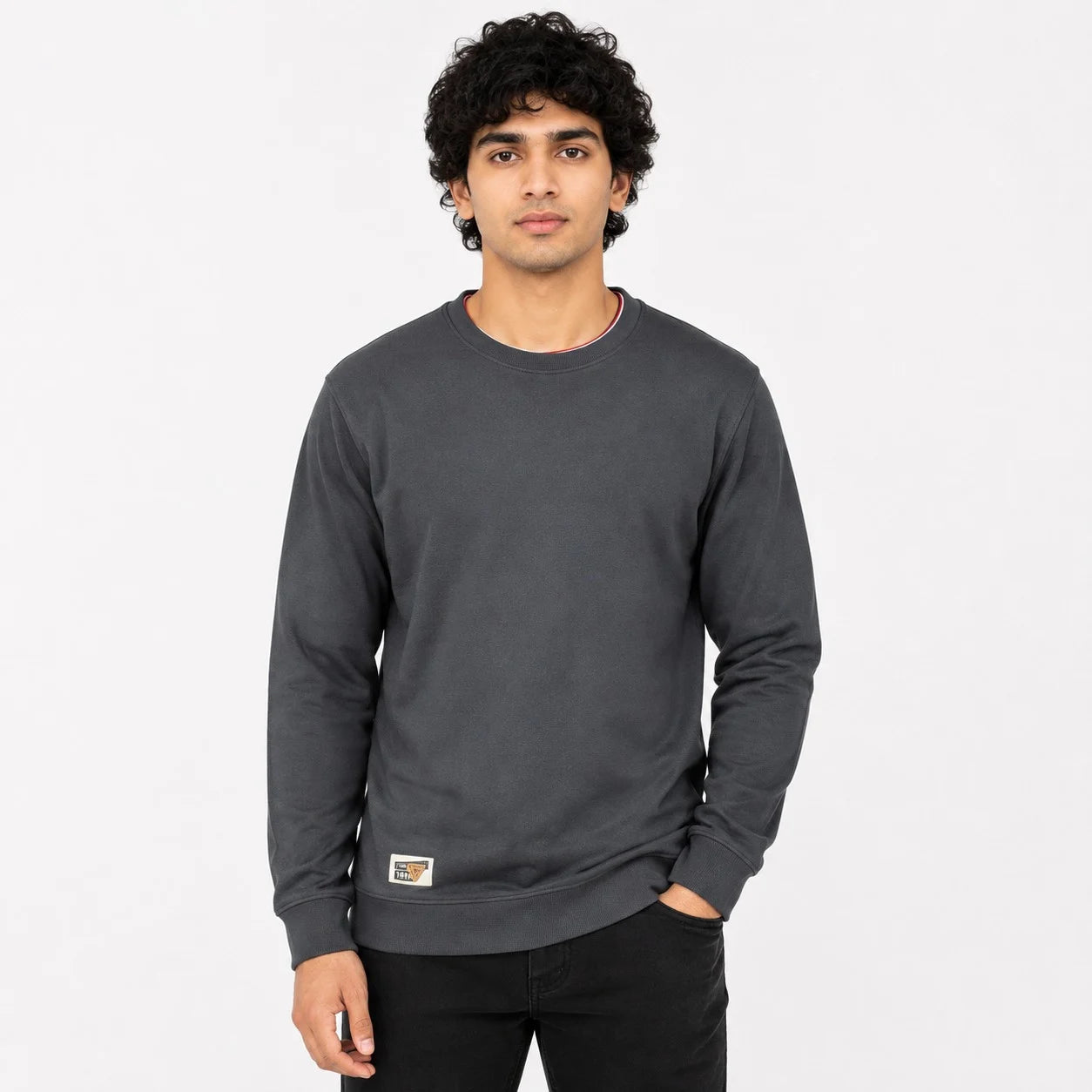 A man with curly black hair wearing a dark gray crewneck sweatshirt and black pants, standing against a plain white studio background.