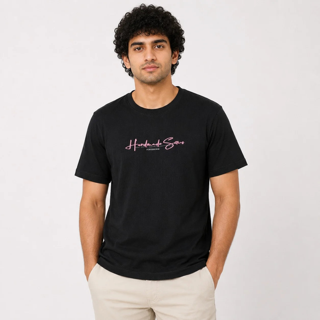 A man wearing a black crew-neck t-shirt with a pink 'Handmade Store' graphic on the chest, paired with beige pants, standing against a light gray background.