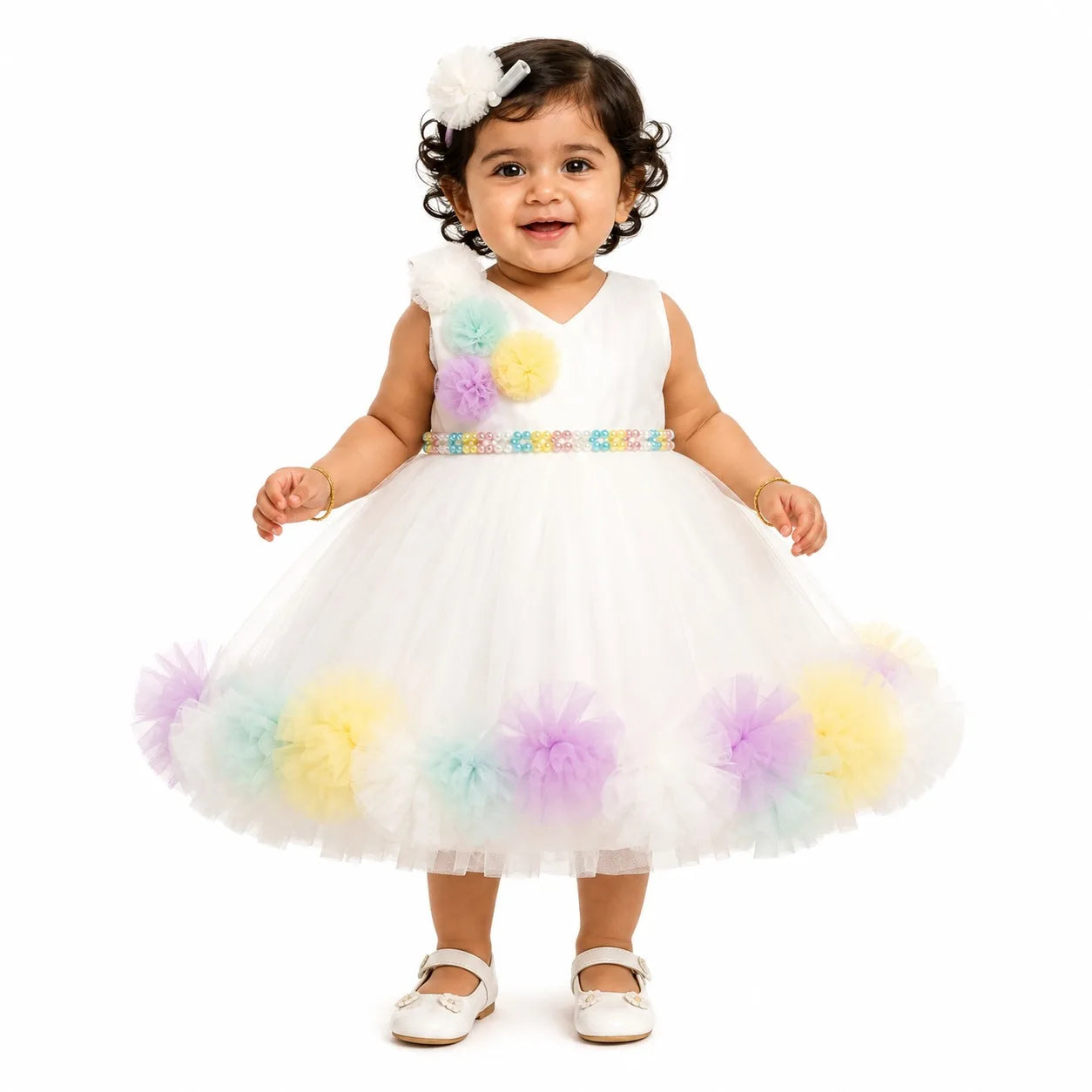 A toddler girl wearing a white sleeveless tutu dress with pastel rainbow pom-poms on the bodice and hem, a matching headband, and white Mary Jane shoes, photographed against a solid white background.