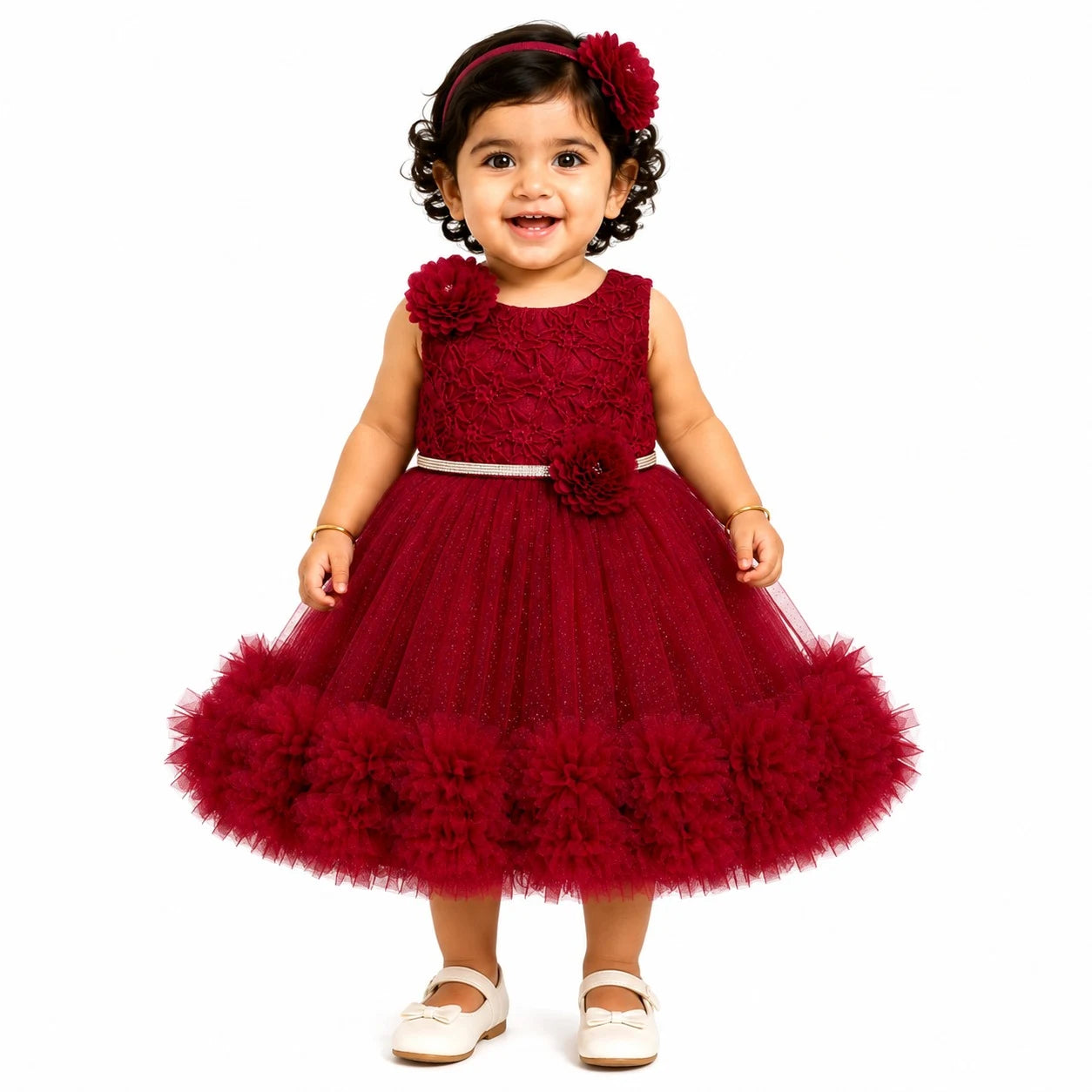 A toddler girl wearing a sleeveless deep red formal party dress with a lace bodice, tulle skirt, and matching red flower headband, standing against a plain white background.