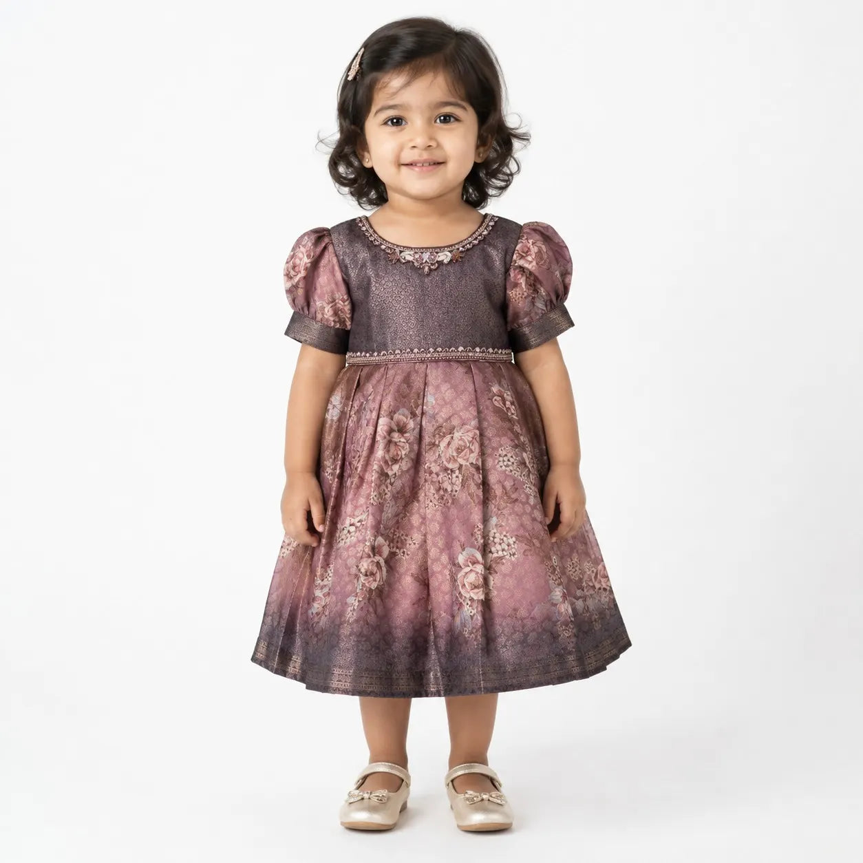 A young toddler girl wearing a short-sleeved dusty rose and purple floral dress with puff sleeves, standing against a plain white studio background.