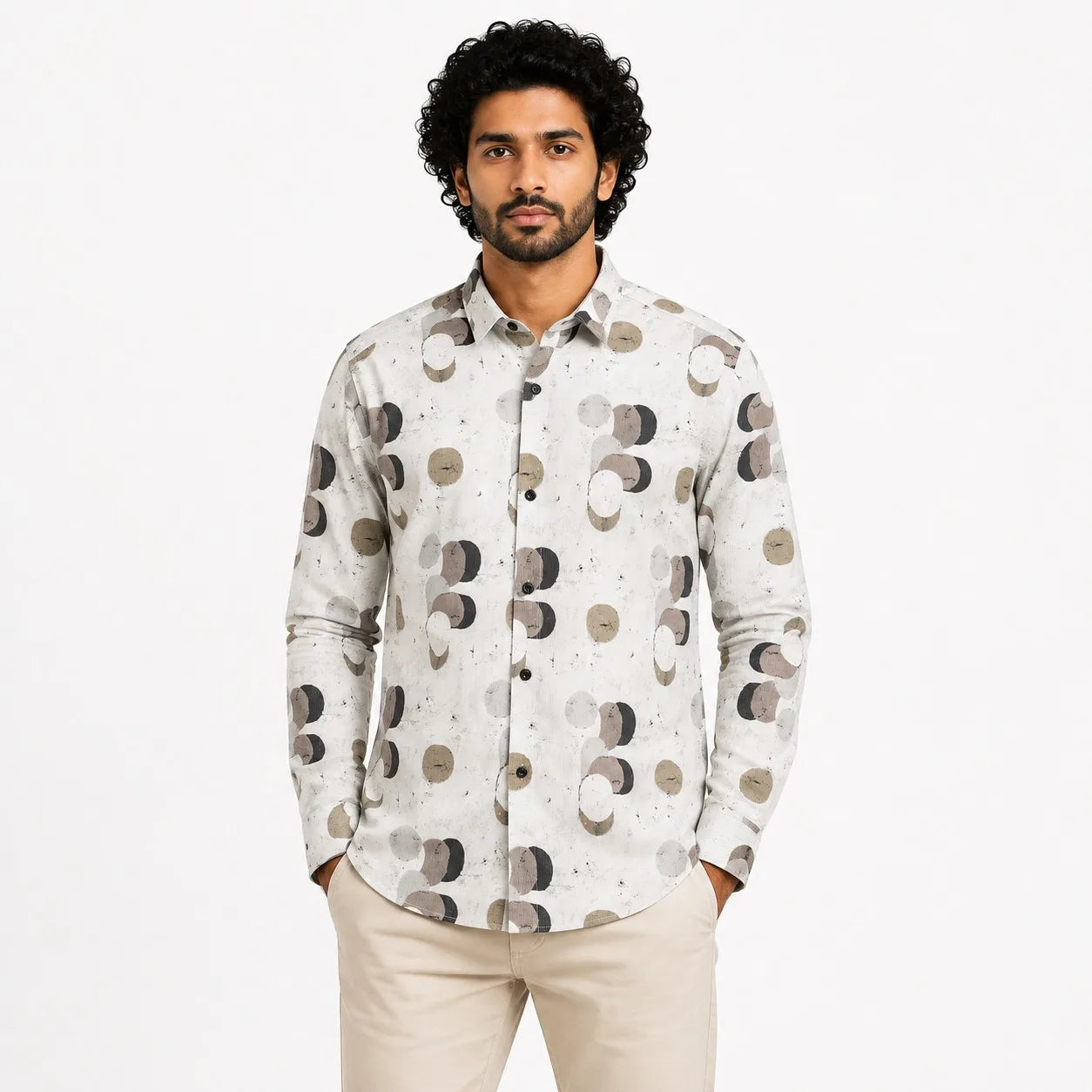 A man wearing a long-sleeve button-up shirt with a white base and a pattern of gray, brown, and beige celestial shapes, paired with beige trousers, against a plain white background.