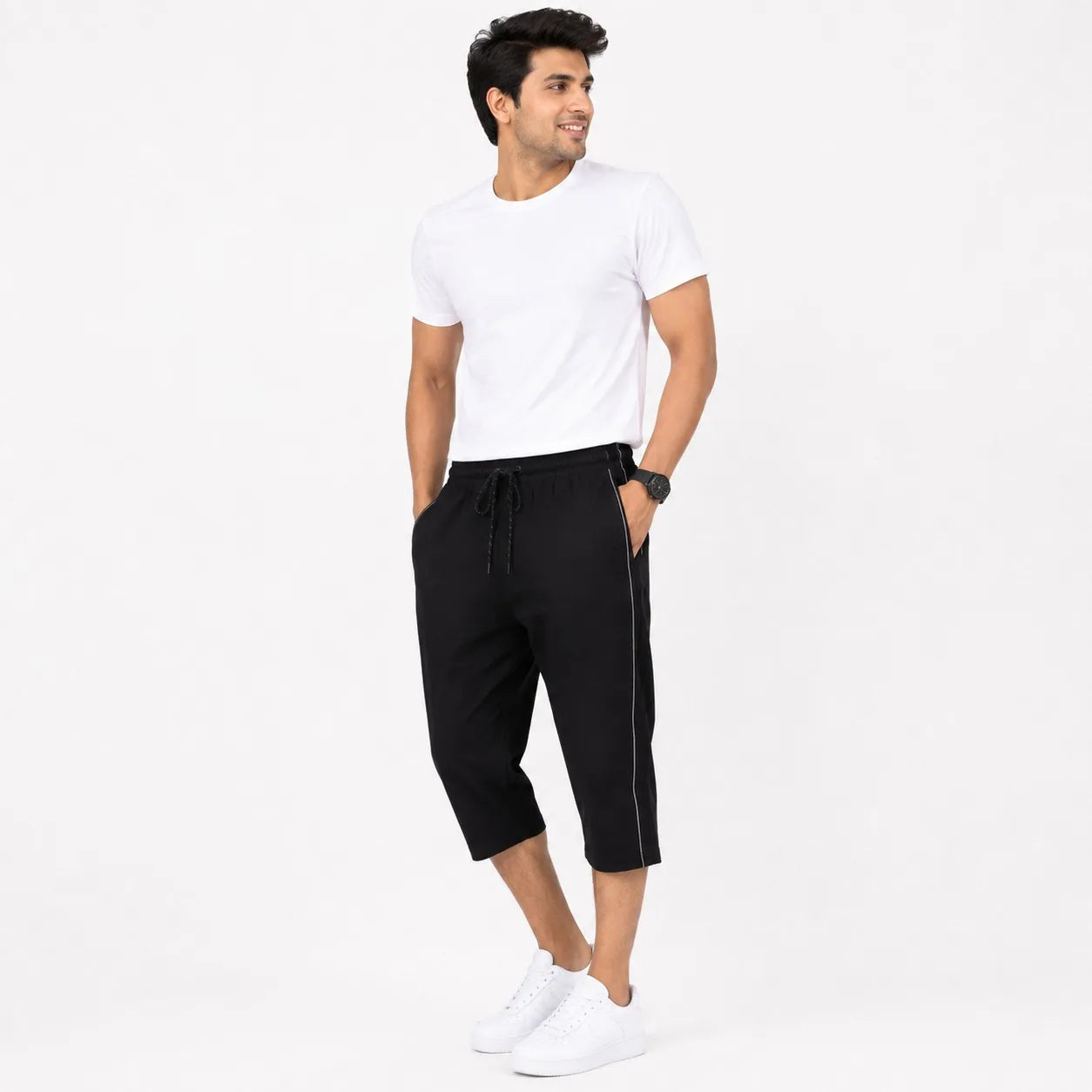 A man modeling black drawstring cropped jogger pants with a thin white side stripe, paired with a white t-shirt and white sneakers, against a plain white background.