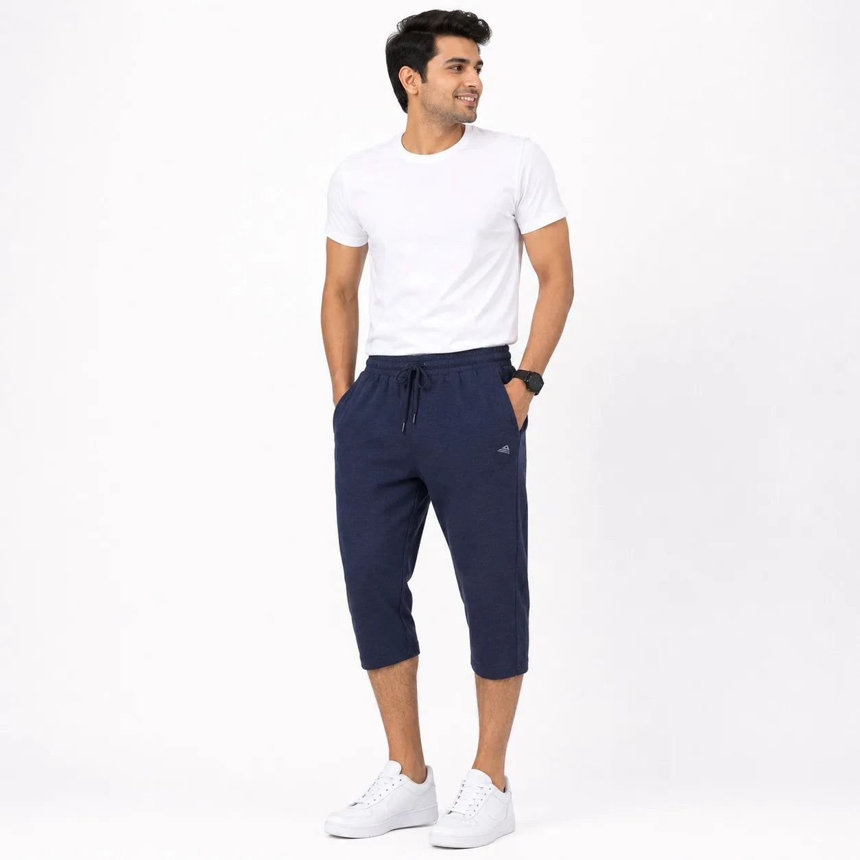 A man wearing a white crew-neck t-shirt, navy blue cropped sweatpants with a drawstring waist, and white sneakers, standing against a plain white studio background.