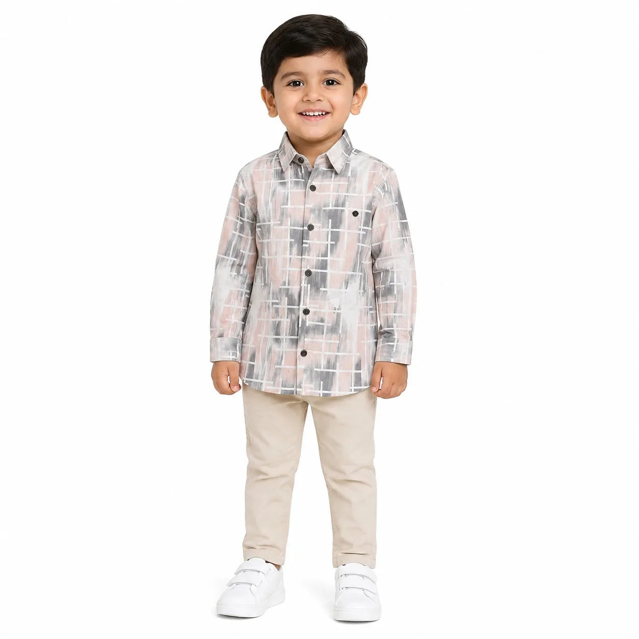 A young boy modeling a long-sleeve button-up shirt with a gray, white, and light pink geometric grid pattern, paired with beige pants and white sneakers, set against a plain white background.