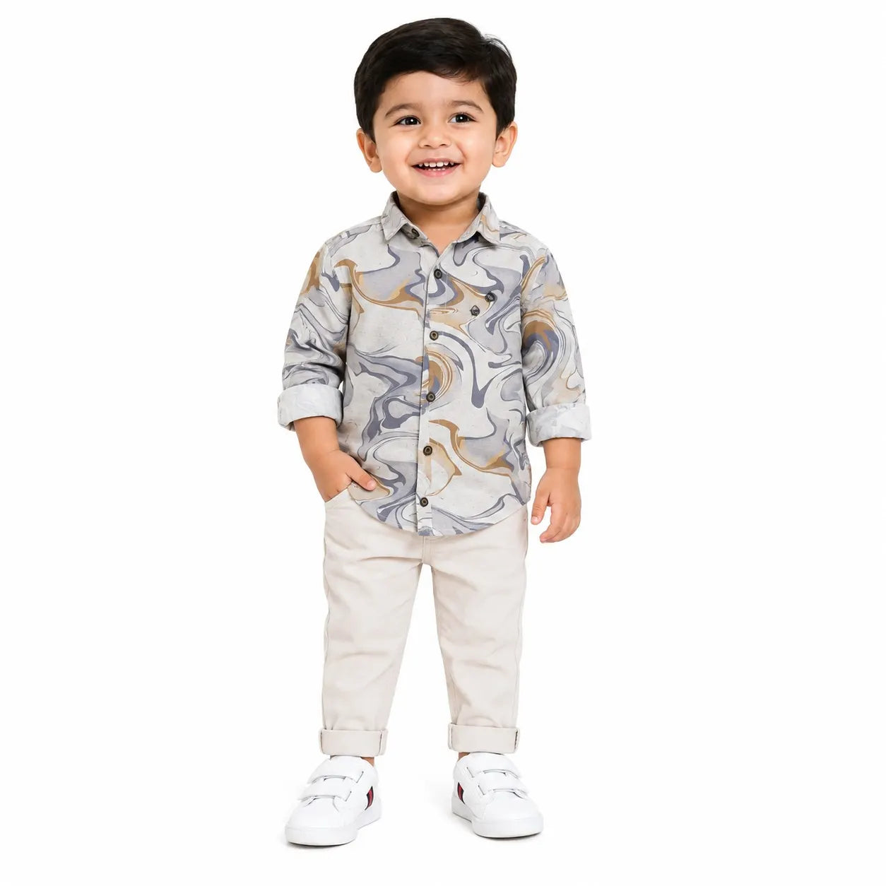 A smiling toddler standing against a plain white background, wearing a long-sleeve button-up shirt with a gray, white, and gold abstract swirl pattern, paired with light beige pants and white sneakers.