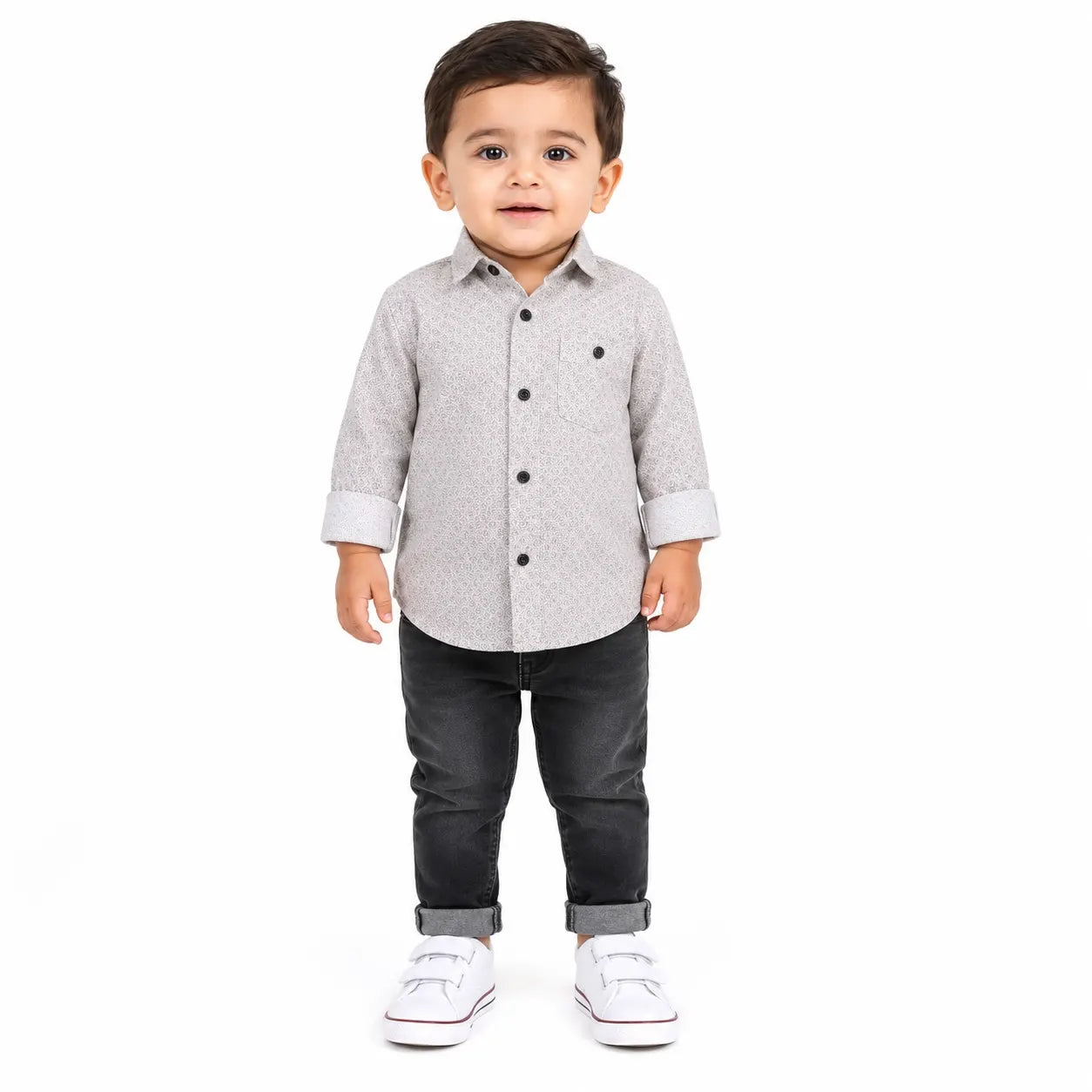 A toddler boy standing against a white background, wearing a light gray patterned long-sleeve button-up shirt, dark gray pants, and white sneakers.
