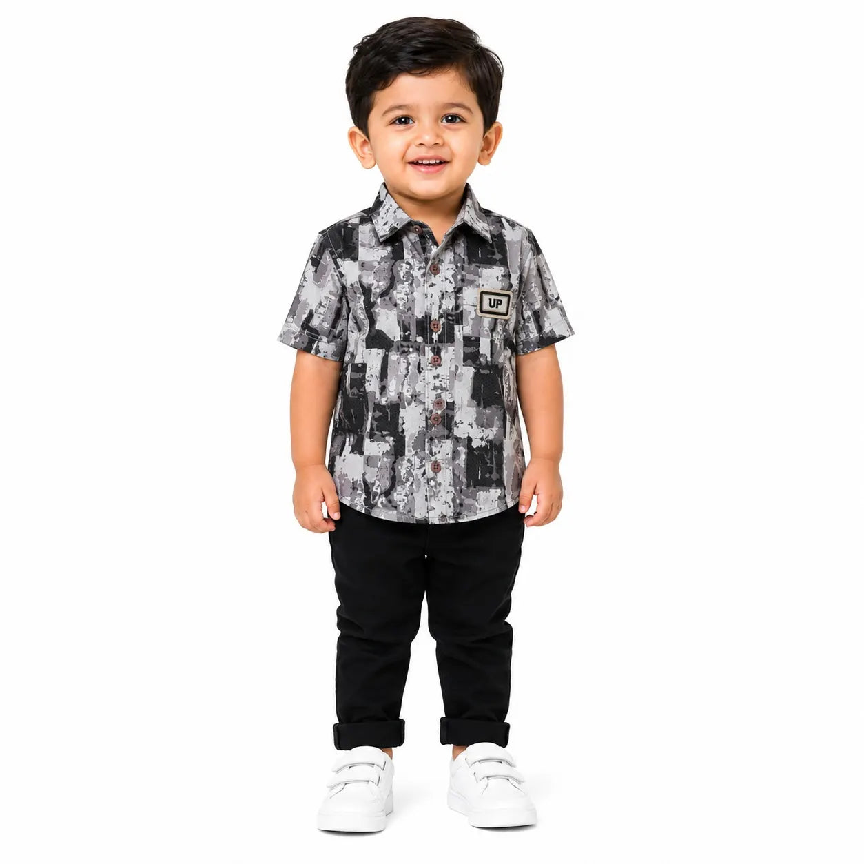 A young boy standing against a white background, wearing a short-sleeve button-up shirt with a black, white, and gray abstract pattern, black pants, and white sneakers.